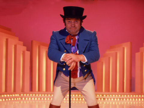 Photograph of Les Dawson performing in a blue jacket against a pink, orange and yellow background on stage.
